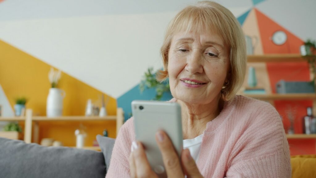 Senhora idosa sorrindo enquanto olha a tela do celular, sentada no sofá de casa.