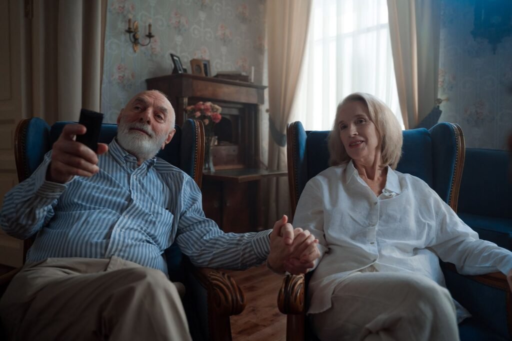 Casal de idosos sentado em poltronas azuis, de mãos dadas, assistindo TV com o controle remoto na mão.